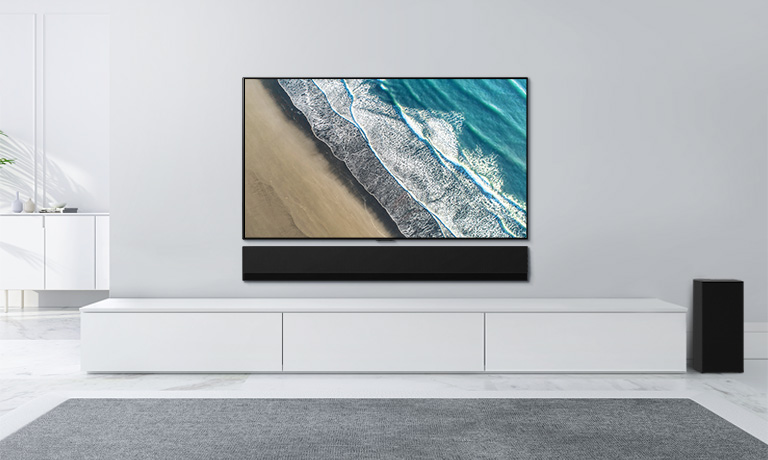 Ein Fernseher ist an einer hellgrauen Wand befestigt. Darunter befindet sich die LG Soundbar auf einer weißen Kommode. Auf dem Fernseher ist ein Strand zu sehen.