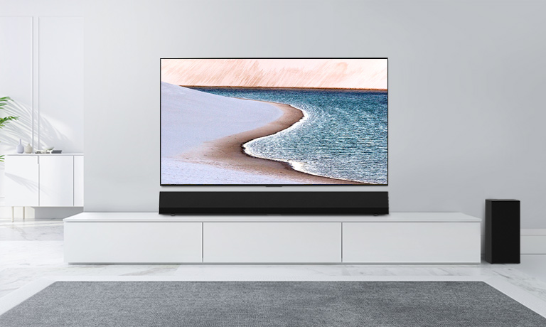 Ein Fernseher ist an einer hellgrauen Wand befestigt. Darunter befindet sich die LG Soundbar auf einer weißen Kommode. Auf dem Fernseher ist ein Strand zu sehen.