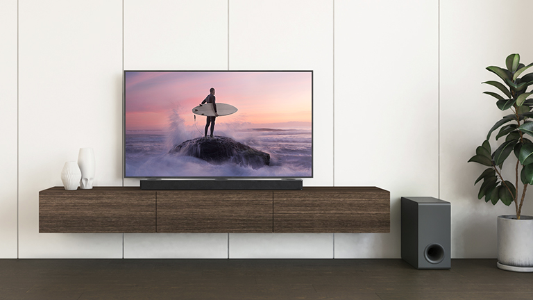 Ein LG TV und eine LG Soundbar stehen auf einem braunen Regal, und der Subwoofer steht auf dem Boden. Der Fernsehbildschirm zeigt einen Surfer, der auf dem Felsen steht.