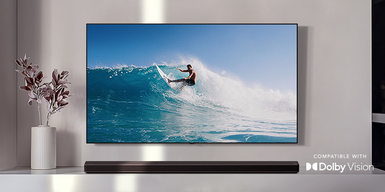 An der Wand befestigter TV. Der TV zeigt einen Mann, der auf einer großen Welle surft. Die LG Soundbar steht direkt unter dem TV auf einem weißen Regal. Direkt neben der Soundbar befindet sich eine Vase mit einer Blume (Video abspielen).