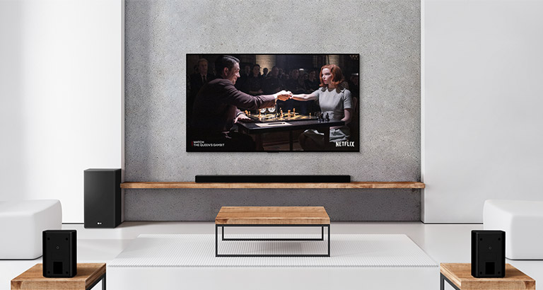 Eine Zusammenstellung, bestehend aus zwei Rücklautsprechern, Subwoofer, einer Soundbar sowie einem TV, befindet sich in einem hellen Wohnzimmer. Ein Poster einer TV-Sendung ist auf dem Bildschirm zu sehen.