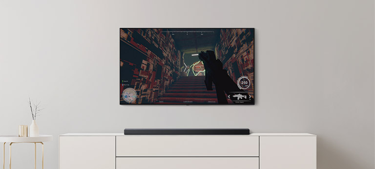 In einem Wohnzimmer befinden sich ein TV und eine Soundbar. Auf dem TV-Bildschirm läuft ein FPS-Spiel und der TV-Kanal ist auf Fußballspiel eingestellt (Video abspielen).