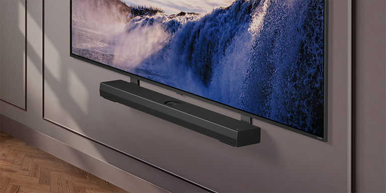 LG-Fernseher erscheint mit einer Synergy-Halterung. Die Synergy-Halterung und der LG-Fernseher sind verbunden. Die Kamera zoomt auf die Synergy-Halterung und zeigt die Soundbar, die auf der Synergy-Halterung platziert ist, gefolgt von dem Hintergrund eines modernen Wohnraums.	