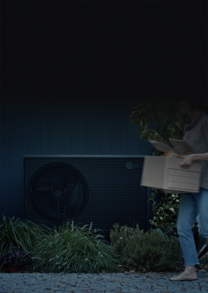 A black LG THERMA V product is placed in the garden of the house and a woman with a box is passing by in front of it.