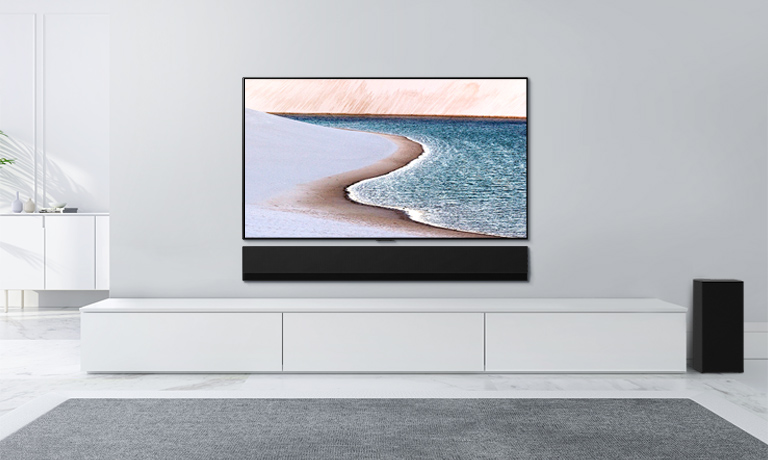 A TV is mounted on light gray wall. LG Soundbar is below it on a white cabinet. The TV shows a beach.