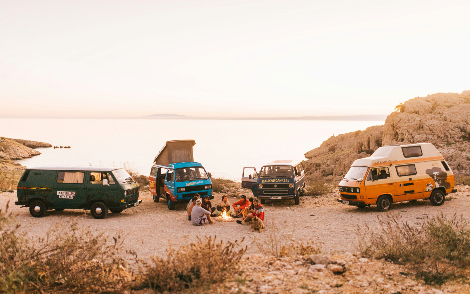 Group of people relaxing by a campfire near three colourful camper vans parked by a seaside at sunset.