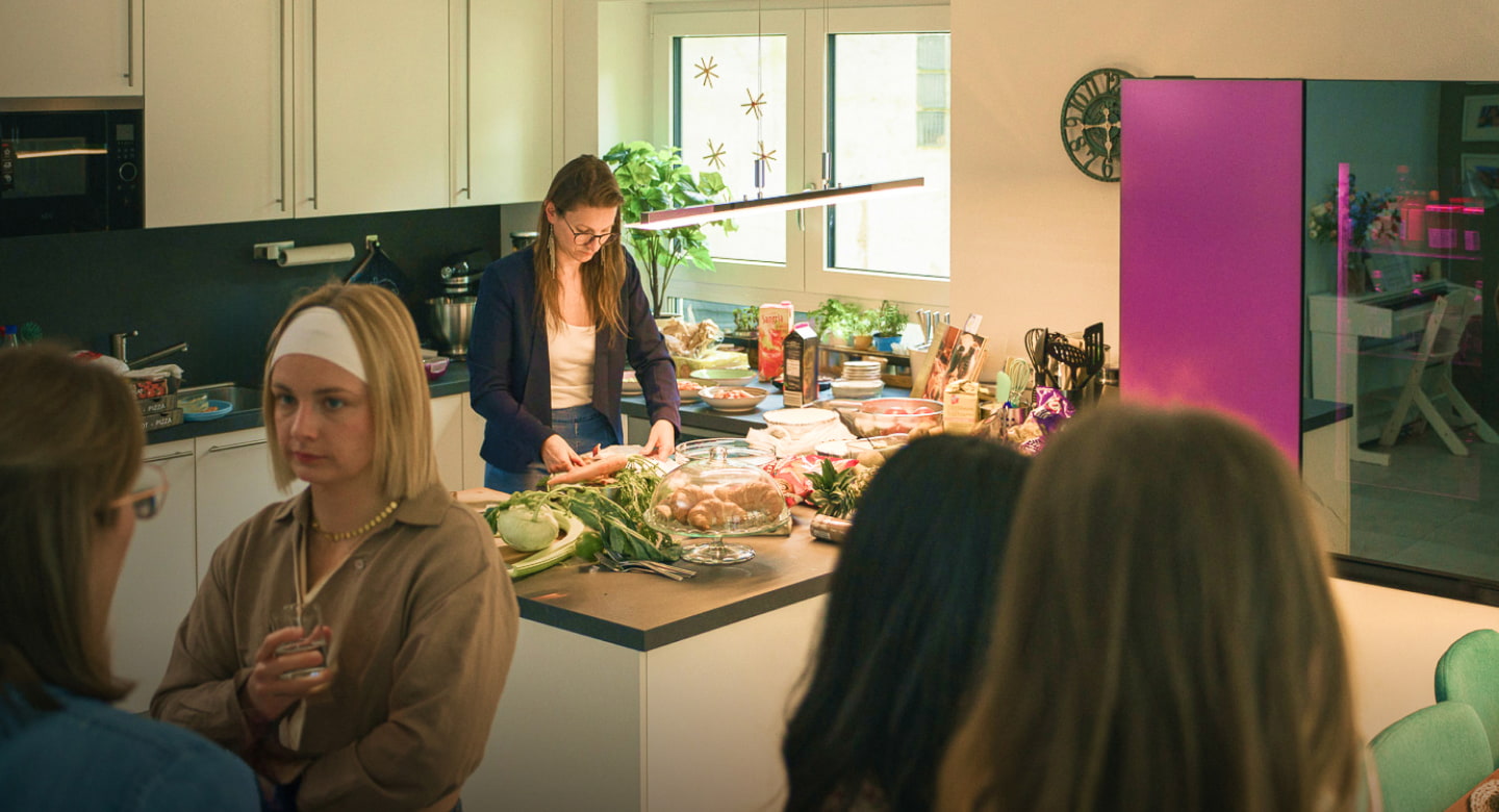 At the home party, friends engage in conversation while the host prepares food at the dining bar.