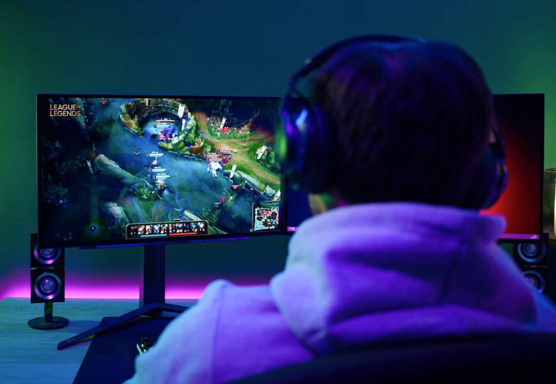 A gamer viewed from the back, playing a game on a dual monitor setup with LED lighting.