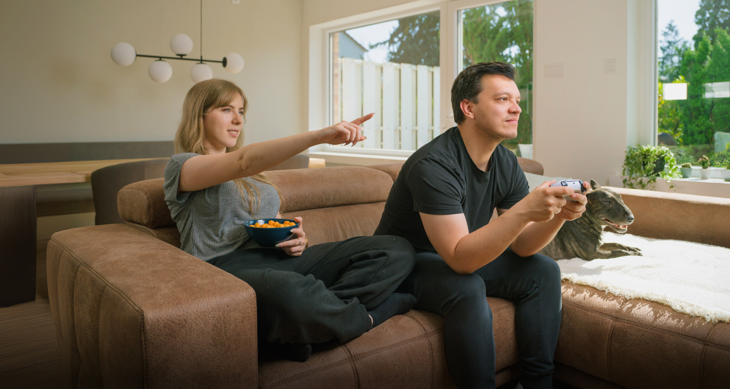 A couple on a couch, with one playing a video game and the other eating snacks.