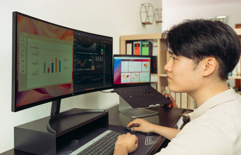 A man working at a modern desk setup with a wide monitors and a laptop on a stand, focusing on charts and data displayed on the screens.