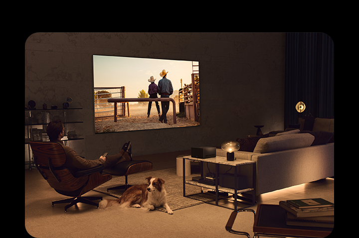 A man watching a cowboy movie with his dog in a cozy living space in the evening. There are no wires surrounding the wall-mounted LG OLED TV, and the Wireless Connect Box sits neatly on a side table. And then, the image slides to another image that shows a man watching a cowboy movie with the  LG OLED TV on a floor stand in a cozy and dark living space. There are no wires surrounding the LG OLED TV and its floor stand, and the Wireless Connect Box sits neatly on a side table.