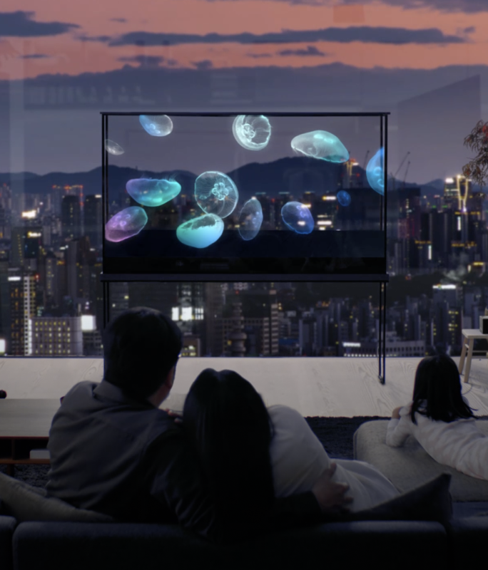 A family enjoys an LG Oled T with a jellyfish floating on it, while the cityscape shines through the transparent screen. 