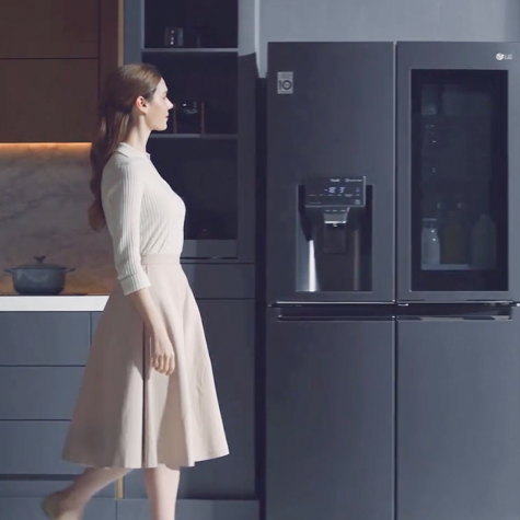A woman walks towards the LG Dios refrigerator and takes out a drink.